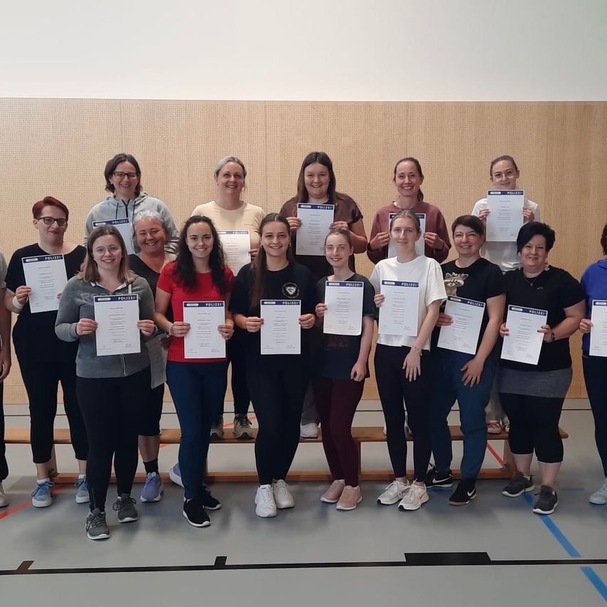 Eine Gruppe von Frauen, wahrscheinlich Kolleginnen oder Freundinnen, hält Urkunden in der Hand und posiert für ein Foto in einer Turnhalle.