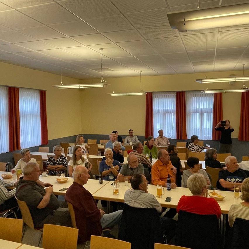 Eine Gruppe von Menschen sitzt an Tischen in einem Raum, wobei eine Person im Hintergrund ein Foto macht. Der Raum hat eine gelbe Wand, rote Vorhänge und eine Decke mit Lichtern.