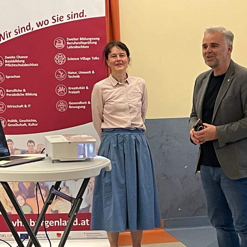 Zwei Personen stehen neben einem Beamer. Eine Frau in einem weißen Hemd und einer blauen Rock lächelt. Ein Mann mit einem grauen Jackett hält ein Handy. Ein Banner mit Symbolen und Text ist hinter ihnen.