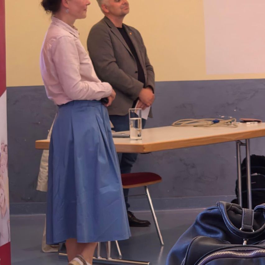 Eine Frau in einem blauen Rock und ein Mann in einem grauen Anzug stehen bei einem Tisch mit einem Glas Wasser.
