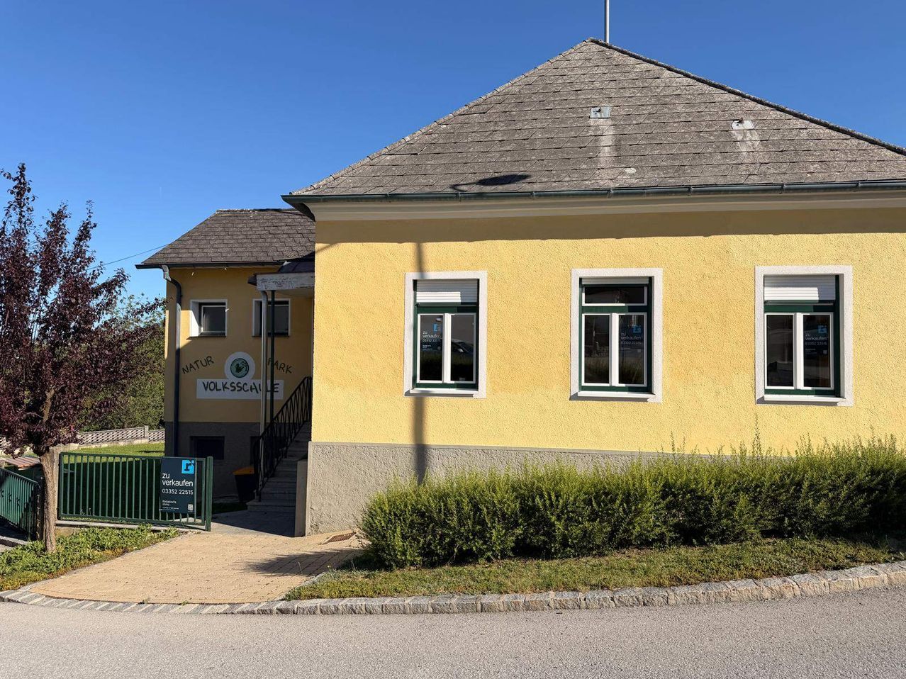 Ein gelbes Gebäude mit einem braunen Dach und grünen Akzenten. Das Gebäude hat ein Schild mit der Aufschrift 'Natur Volksschule' und einen Eingang mit Treppen.