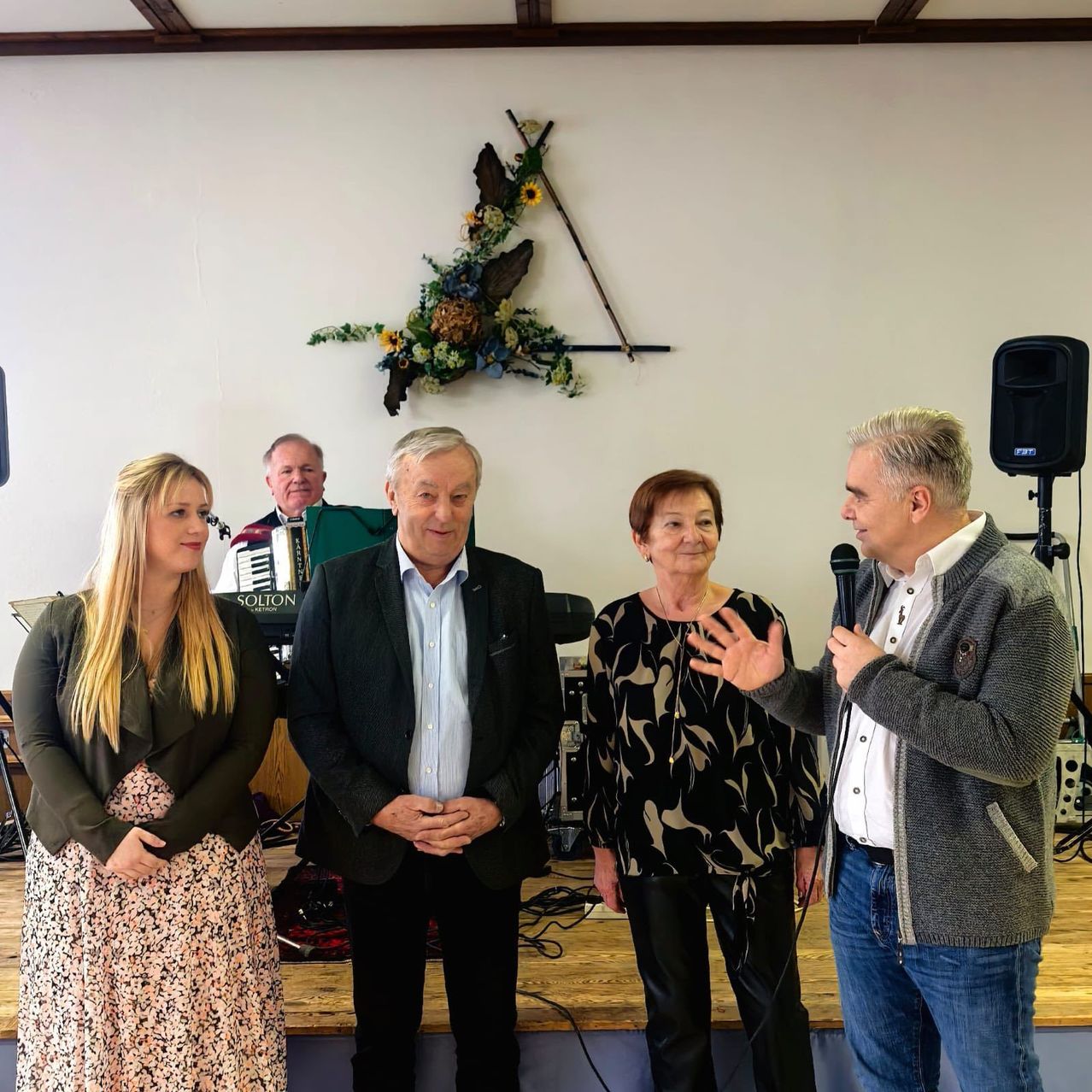 Eine Gruppe von Menschen steht auf einer Bühne mit einem Lautsprecher. Ein Mann hält ein Mikrofon und spricht. Hinter ihnen ist eine Blumendekoration und ein Mann, der Akkordeon spielt.