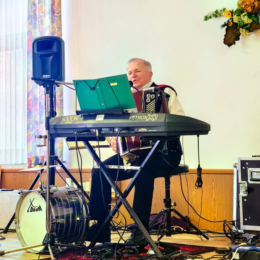 Ein älterer Mann spielt Akkordeon, während er auf einem Hocker in einem Raum mit Schlagzeug und Lautsprecher auf dem Boden sitzt.
