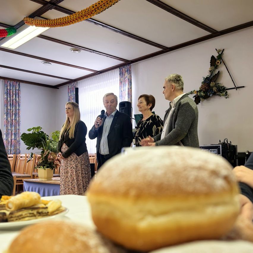 Eine Gruppe von Menschen steht in einem Raum mit einem Tisch voller Brot und Gebäck. Ein Mann mit einem Mikrofon spricht, während eine Frau in der Nähe steht. Ein Lautsprecher und eine Pflanze sind im Hintergrund.