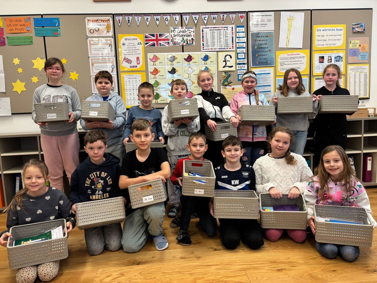 Ein Klassenfoto von Kindern, die Aufbewahrungsboxen halten, mit Dekorationen an der Wand, darunter Alphabet-Charts, Zahlen und eine britische Flagge.