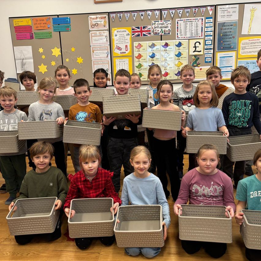 Eine Gruppe von Kindern in einem Klassenzimmer, alle graue Aufbewahrungsboxen haltend, lächeln für ein Foto. Das Klassenzimmer hat farbenfrohe Poster und eine Pinnwand mit verschiedenen Diagrammen und Informationen.