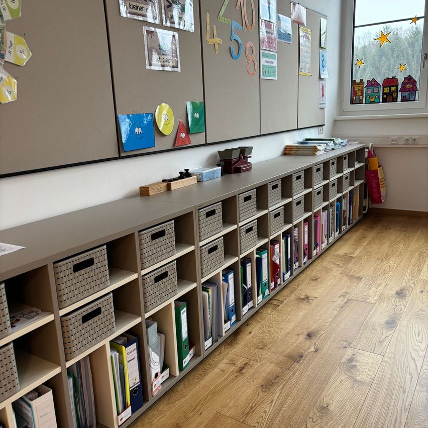 Ein Klassenzimmer mit Holzboden, grauen Regalen und Büchern. Die Wand hat farbenfrohe Dekorationen und Papiere.