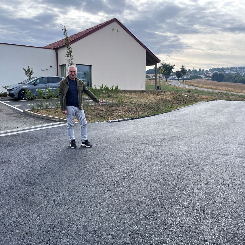 Ein Mann in einer Jacke und Turnschuhen steht vor einer frisch asphaltierten Straße, lächelt und zeigt. Hinter ihm steht ein modernes Gebäude mit einem geparkten Auto davor.