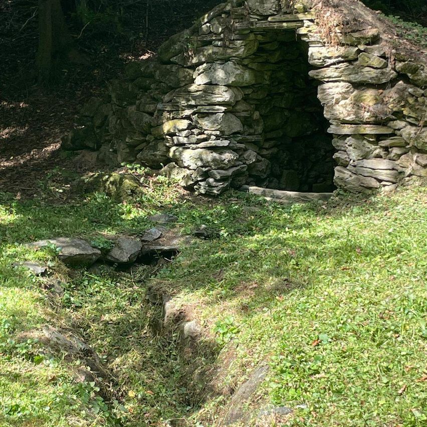 Eine kleine Steinstruktur mit einem Eingang in einem Grasbereich, umgeben von Felsen und Moos.
