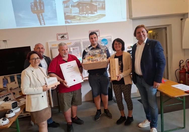 Eine Gruppe von Menschen steht in einem Raum, lächelt und hält ein Zertifikat und einen Geschenkkorb.