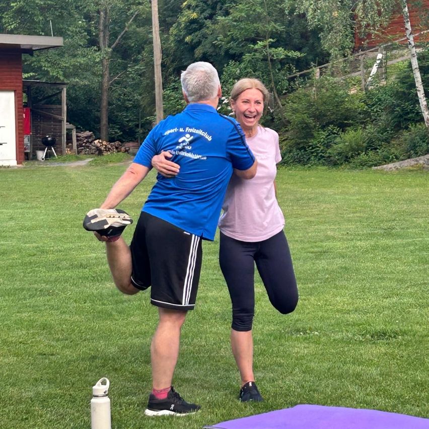 Ein Mann und eine Frau lachen und umarmen sich auf einer Wiese, der Mann streckt sein Bein. Eine Flasche und eine Yogamatte sind in der Nähe. Hinter ihnen sind ein Haus und ein Holzschuppen zu sehen.