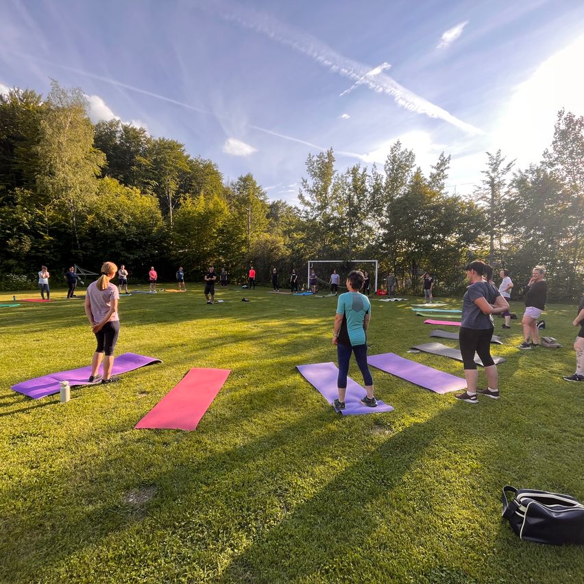 Eine Gruppe von Menschen macht Yoga auf einem Feld mit grünem Gras. Einige von ihnen stehen, während andere auf Yogamatten sitzen. Das Gebiet ist von Bäumen umgeben und hat ein Fußballtor. Der Himmel ist blau mit einigen Wolken.