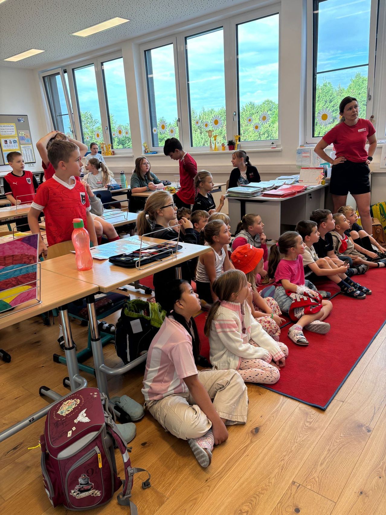 Ein Klassenzimmer mit Kindern auf dem Boden sitzend, eine Lehrerin vor ihnen stehend und Tische und Stühle im Hintergrund angeordnet.