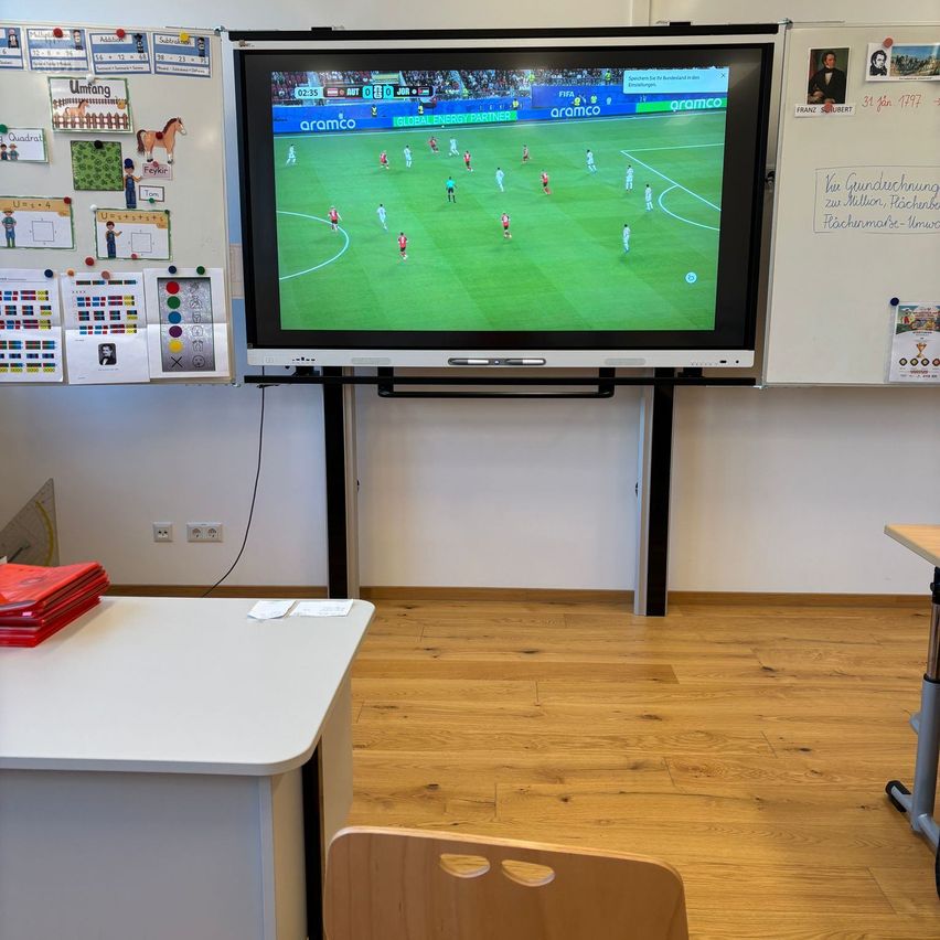 Ein Klassenzimmer mit einem großen Monitor, der ein Fußballspiel zeigt. Hinter dem Monitor befindet sich eine Tafel mit verschiedenen Lernmaterialien. Ein Schreibtisch mit Büchern und ein Holzstuhl sind im Vordergrund.