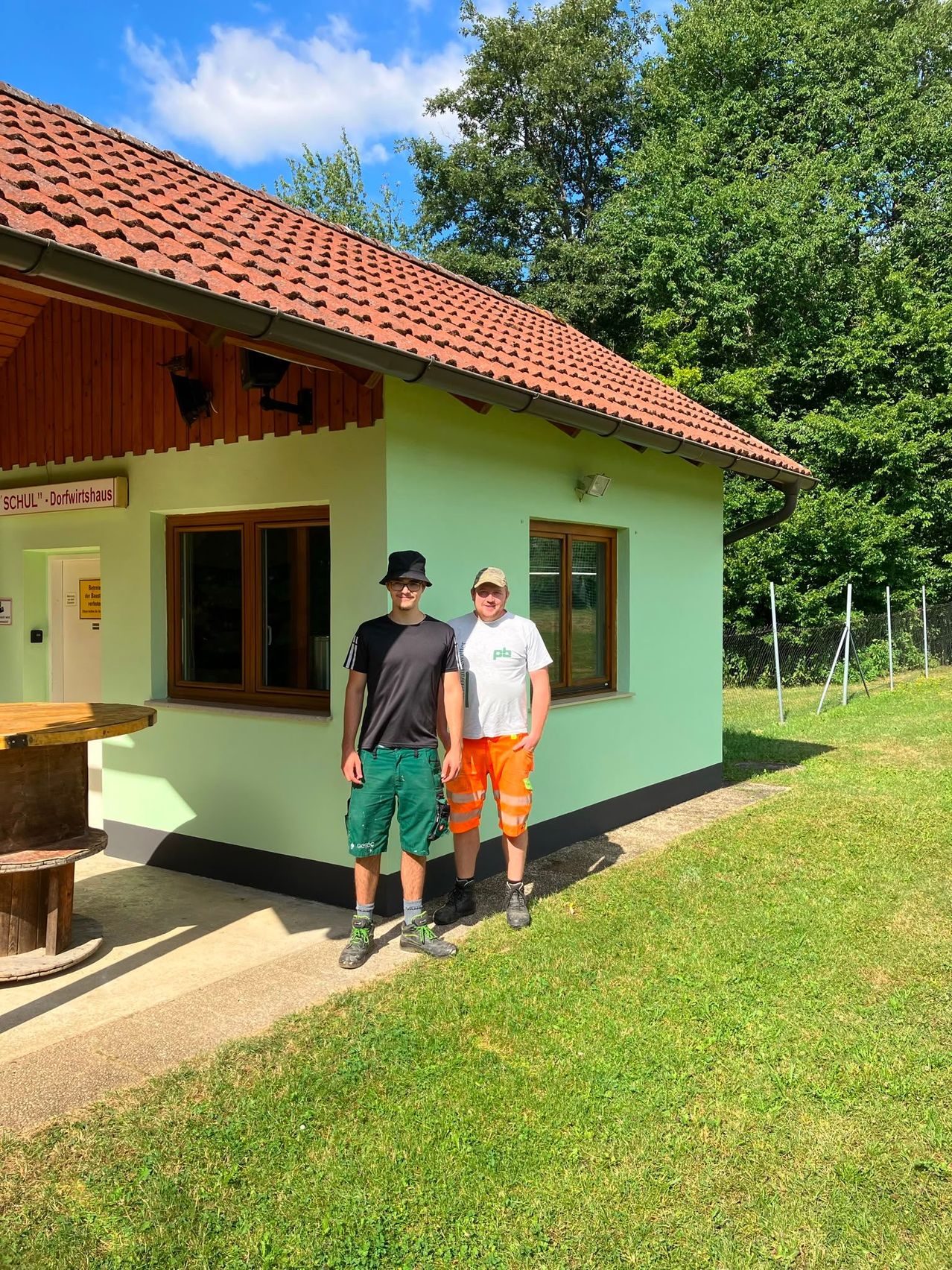 Zwei Männer stehen vor einem kleinen Gebäude mit rotem Dach und grünen Wänden. Sie tragen beide Mützen und Turnschuhe. Das Gebäude hat ein Schild, das 'SCHUL-Dorfthaus' besagt.