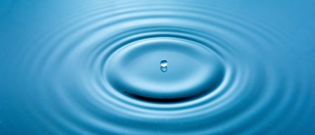 Ein Wassertropfen ist in einen ruhigen, blauen Wasserkörper gefallen und hat dabei Wellen erzeugt, die sich nach außen ausbreiten.