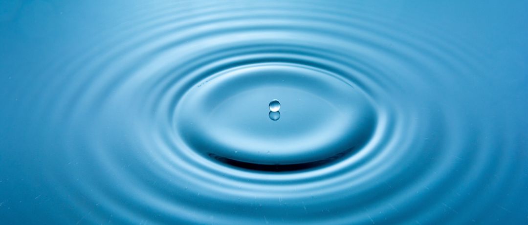 Ein Wassertropfen ist in einen ruhigen, blauen Wasserkörper gefallen und hat dabei Wellen erzeugt, die sich nach außen ausbreiten.