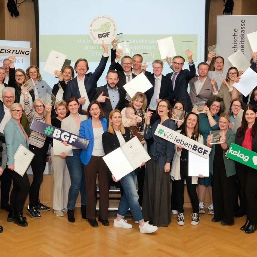 Eine Gruppe von Menschen posiert für ein Foto, hält Papiere und Urkunden in den Händen, während einige die Hände heben. Ein Mann in der Mitte hält ein Papier mit dem Text BGF. Hinter ihnen ist eine Projektionsleinwand an der Wand befestigt.