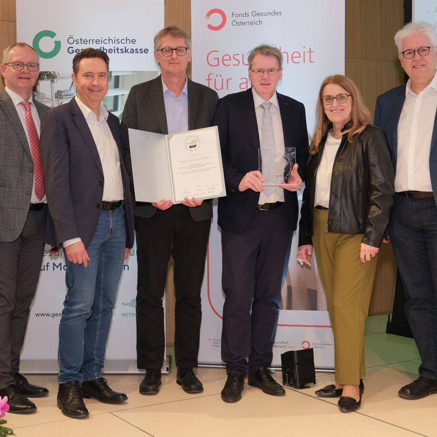 Eine Gruppe von Personen steht in formeller Kleidung und posiert für ein Foto vor einem Banner. Zwei Männer halten Auszeichnungen, andere lächeln. Im Hintergrund ist ein Logo und Text zu sehen.