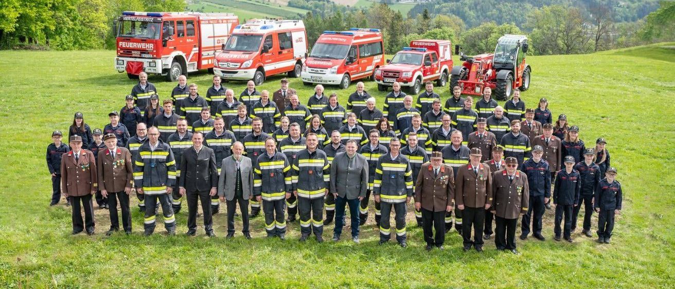 Eine große Gruppe von Feuerwehrleuten und Beamten posiert für ein Foto vor mehreren Feuerwehrwagen und einem Traktor auf einer Wiese mit einer malerischen Kulisse.