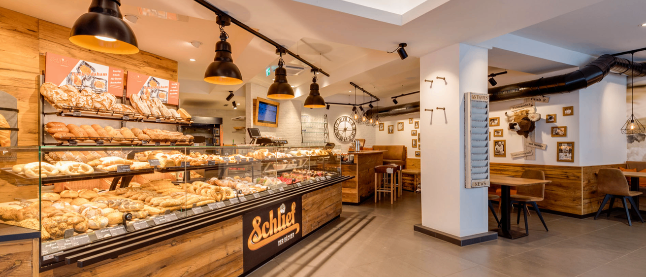 Innenansicht einer Bäckerei mit einer Glasvitrine voller verschiedener Gebäckstücke, an der Decke hängende Pendelleuchten, eine Holztheke und eine Uhr an der Wand.