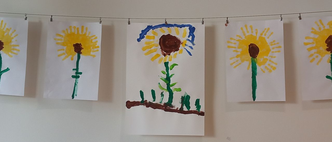 Eine handgemalte Sonnenblume auf weißem Papier hängt an einer weißen Wand mit anderen Gemälden.