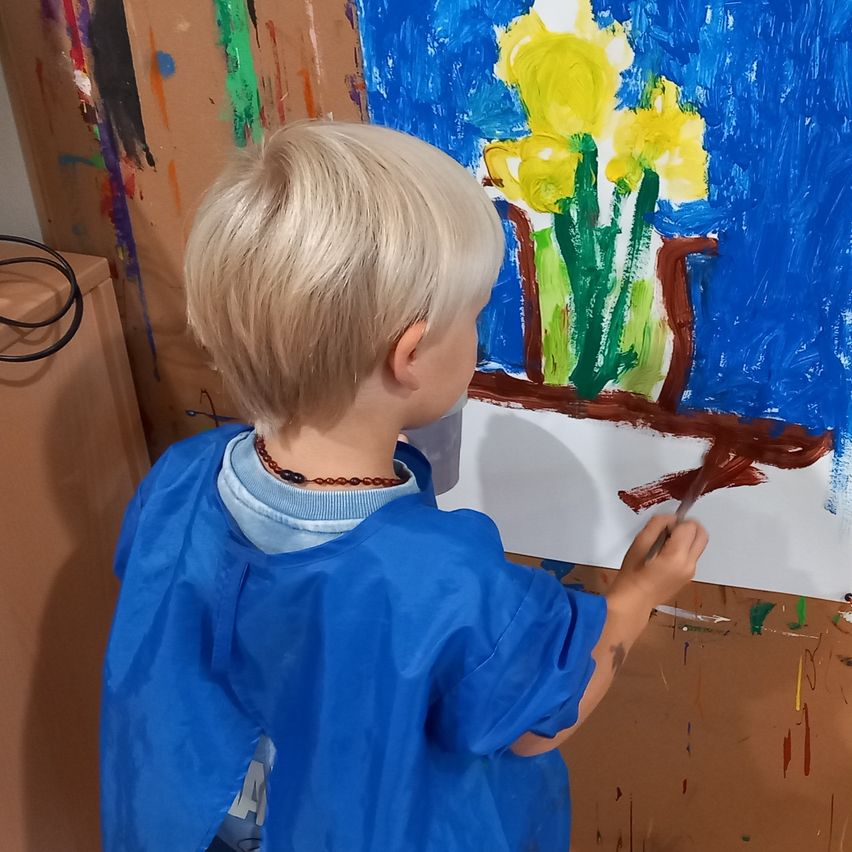 Ein junger Junge malt mit einem Pinsel auf eine Leinwand. Er trägt einen blauen Kittel. Das Gemälde zeigt einen Strauß gelber Blumen in einem blauen Hintergrund.