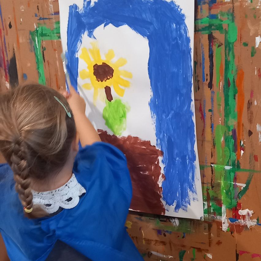 Ein junges Mädchen malt eine Sonnenblume auf eine große Leinwand. Sie trägt ein blaues Hemd und einen weißen Spitzenkragen. Die Leinwand ist an einer Wand mit verschiedenen Farbspritzern montiert.