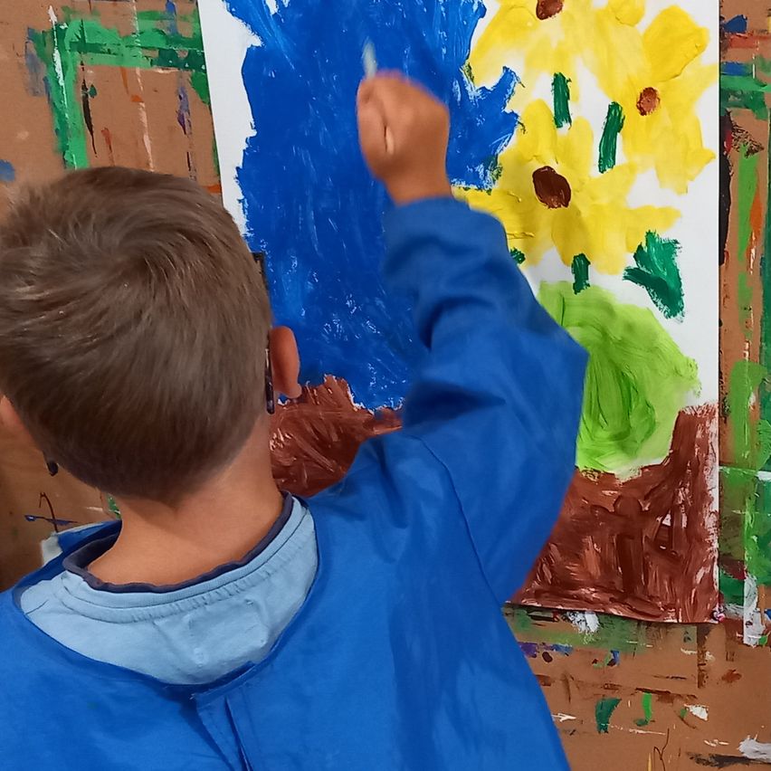 Ein junger Junge in einem blauen Malkittel malt ein Bild mit blauen und gelben Farben auf einem braunen Hintergrund.