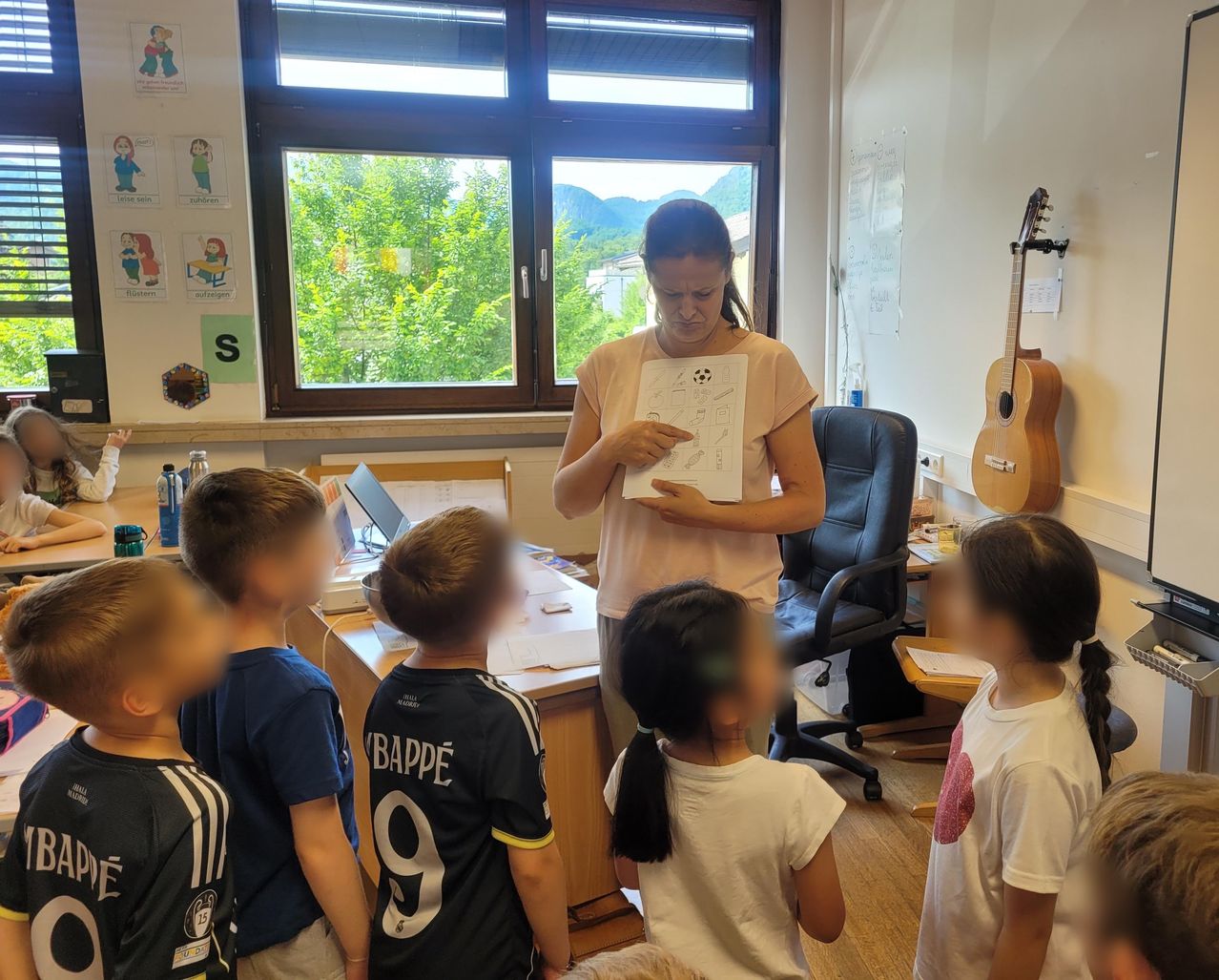 Eine Frau steht in einem Klassenzimmer und zeigt Kindern ein Papier. Der Raum hat ein Fenster mit Blick auf Bäume und Berge. Verschiedene Poster und eine Gitarre sind sichtbar.