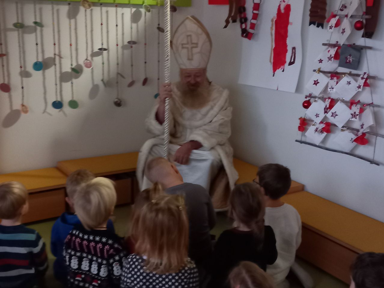 Ein Priester in einer weißen Robe und Mitra spricht eine Gruppe von Kindern an, die auf dem Boden in einem Klassenzimmer sitzen.