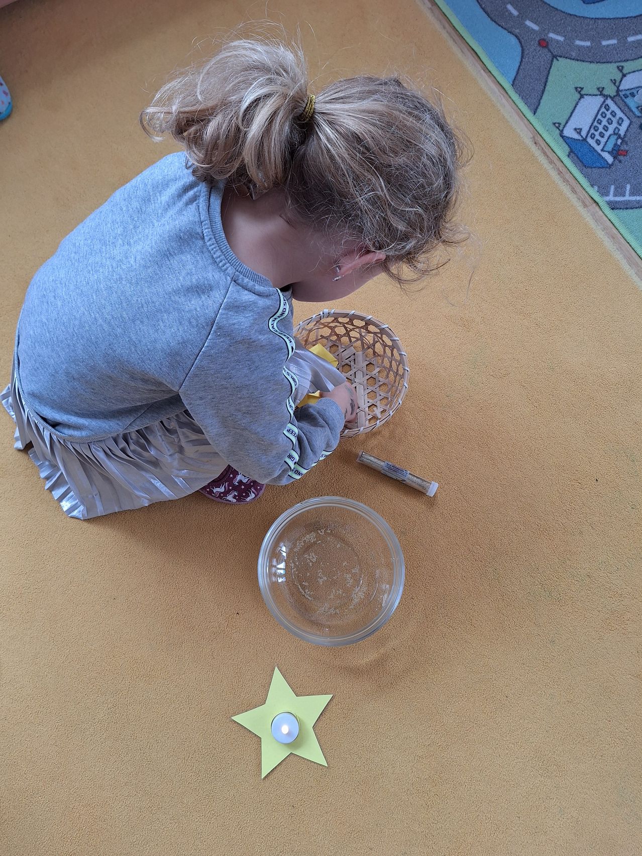 Ein Mädchen in einem grauen Shirt und einer Rock sitzt auf dem Boden und schaut in einen Korb. Der Korb ist mit gelben Objekten gefüllt. Es gibt eine Schüssel mit Wasser und eine Kerze in einem sternförmigen Papier in der Nähe.