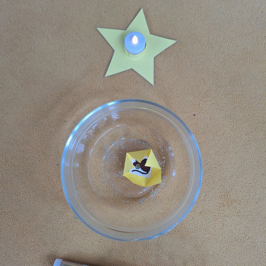 Ein mit Wasser gefüllter Behälter enthält ein gelbes Papierboot mit einer Katze darauf. Ein grüner Stern aus Papier und eine Kerze befinden sich neben dem Behälter auf dem Teppich.