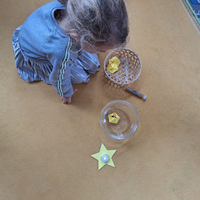 Ein kleines Mädchen kniet auf dem Boden und schaut in einen Korb mit gelben Papierbooten. Ein gelber Stern und ein kleines Licht sind auf dem Boden.