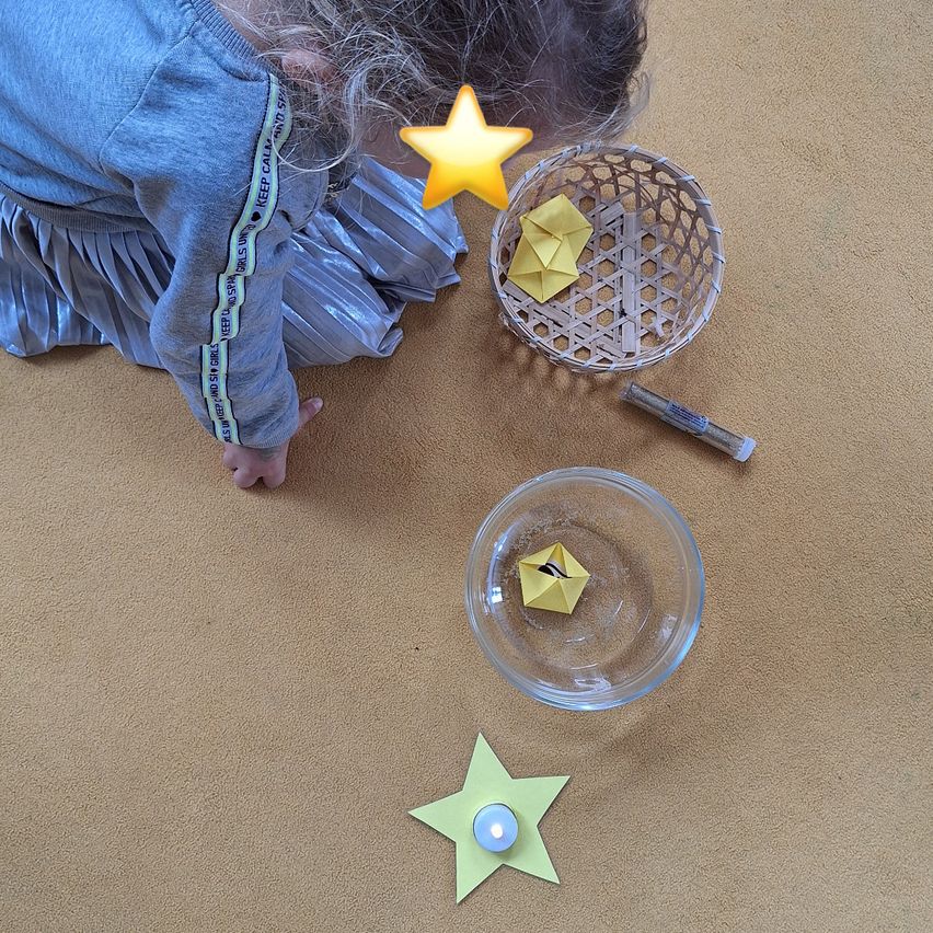 Ein Kind kniet auf einem Teppichboden, umgeben von einer Schüssel, einem Korb und Sternen. Das Kind hält einen Stern und schaut auf einen gelben Papierstern in einer Schüssel.