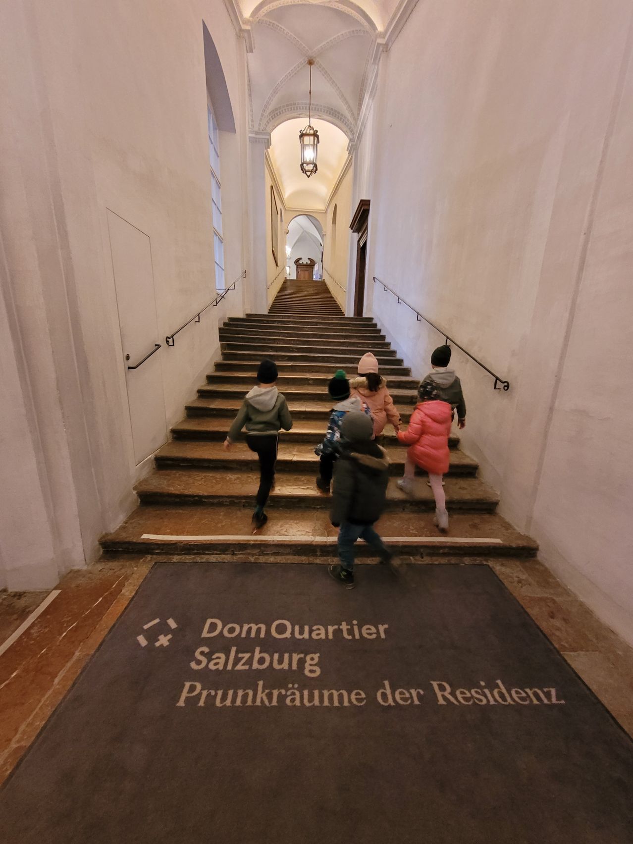 A group of children are running up a staircase in an old building. The staircase is lined with handrails and leads to a corridor. The floor is carpeted, and there is a sign that reads Dom Quartier Salzburg Prunkraume der Residenz.