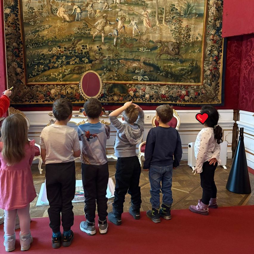 Five children are standing on a red carpet, facing a large tapestry with a historical scene. They appear to be engaged and focused on the artwork.