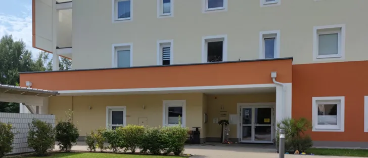 Die Vorderseite eines modernen Wohngebäudes mit orangen und weißen Wänden, ausgestattet mit mehreren Fenstern und einem Doppeleingang. Das Äußere ist von einem Garten mit Sträuchern und Rasen umgeben.