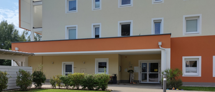 Die Vorderseite eines modernen Wohngebäudes mit orangen und weißen Wänden, ausgestattet mit mehreren Fenstern und einem Doppeleingang. Das Äußere ist von einem Garten mit Sträuchern und Rasen umgeben.