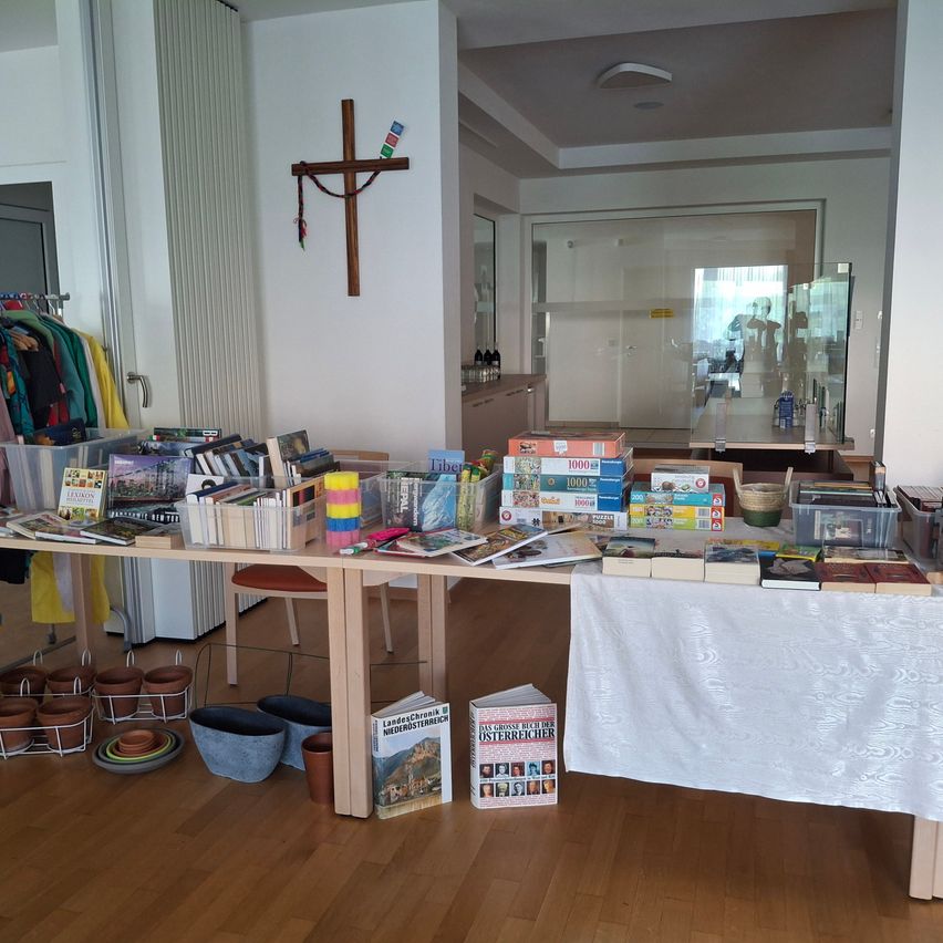 Ein Raum mit einem Tisch voller Bücher, Puzzles und anderen ausgestellten Gegenständen. Ein Kreuz ist an der Wand montiert.