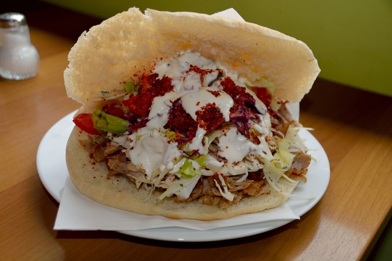 Ein Nahaufnahme eines mit Fleisch gefüllten Pita-Sandwichs mit Tomaten, Salat und weißer Sauce auf einem weißen Teller, auf einem Serviettentuch auf einem Holztisch.