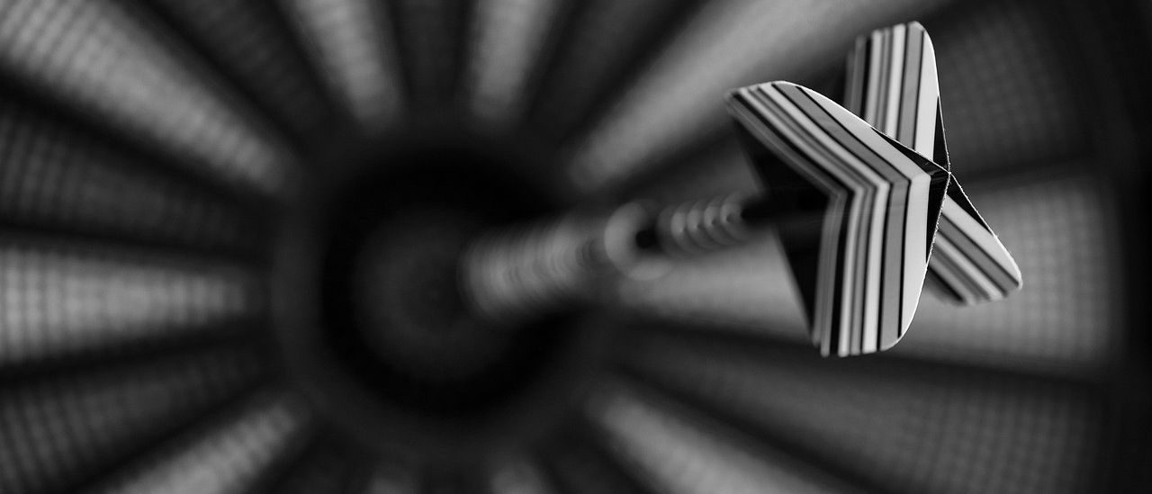 Ein schwarz-weißes Bild zeigt einen im Dartboard steckenden Dart, wobei die Spitze und die Schwanzfedern des Darts sichtbar sind.