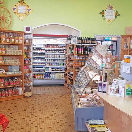 Ein Geschäft mit Holzregalen, die mit verschiedenen Produkten gefüllt sind. Hinter den Regalen befindet sich eine Kühltheke, die eine Auswahl an Lebensmitteln präsentiert. Die Wand ist grün mit dekorativen Gegenständen. Rechts gibt es eine Theke mit Produkten im Schaufenster.