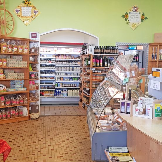 Ein Geschäft mit Holzregalen, die mit verschiedenen Produkten gefüllt sind. Hinter den Regalen befindet sich eine Kühltheke, die eine Auswahl an Lebensmitteln präsentiert. Die Wand ist grün mit dekorativen Gegenständen. Rechts gibt es eine Theke mit Produkten im Schaufenster.