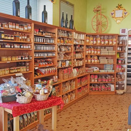 Ein Geschäft mit verschiedenen Waren, darunter Gläser, Flaschen und Körbe, auf Holzregalen und einem Tisch mit roter Tischdecke. Flaschen stehen oben auf den Regalen, und an der Wand befindet sich ein dekoratives Rad.