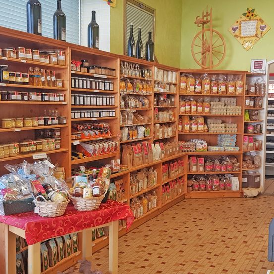 Ein Geschäft mit verschiedenen Waren, darunter Gläser, Flaschen und Körbe, auf Holzregalen und einem Tisch mit roter Tischdecke. Flaschen stehen oben auf den Regalen, und an der Wand befindet sich ein dekoratives Rad.