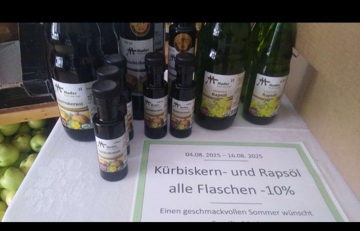 Flaschen mit Kürbiskern- und Rapsöl auf einem Tisch mit einem Schild, das 'Alle Flaschen -10%' anzeigt. Äpfel sind im Hintergrund.