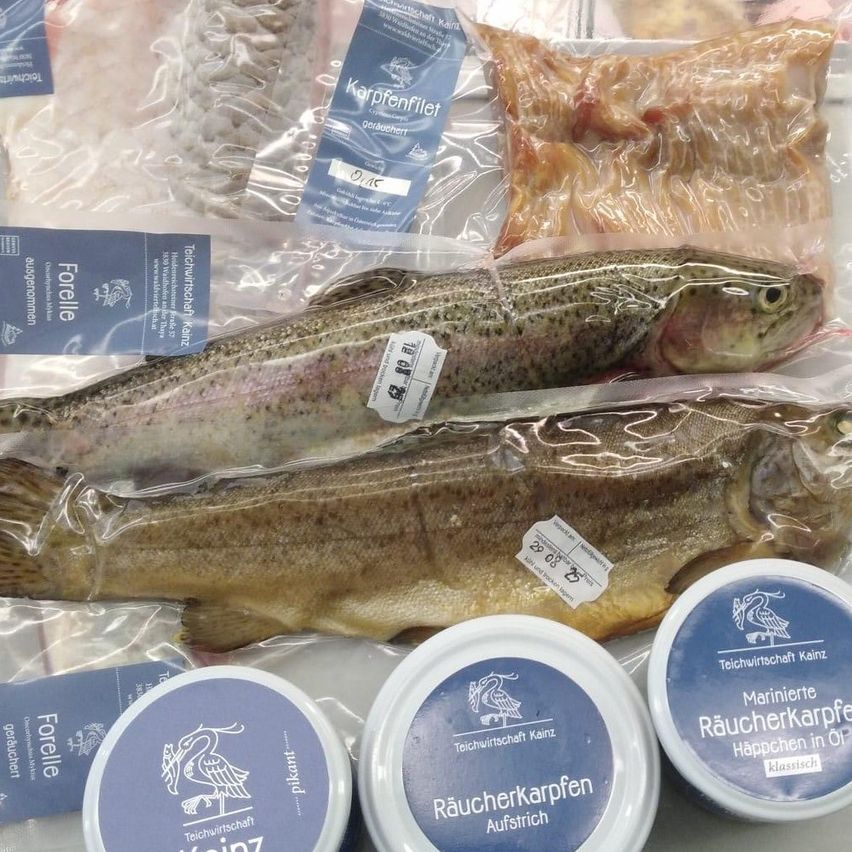 Ein Nahaufnahme von zwei Forellenfischen, zwei Fleischpaketen und drei Gläsern mit geräucherten Fischprodukten wird auf einem blauen Hintergrund gezeigt. Die Forellenfische sind in Plastik verpackt mit Etiketten. Das Fleisch ist ebenfalls in Plastikverpackungen. Die geräucherten Fischprodukte sind in Gläsern mit Deckeln.