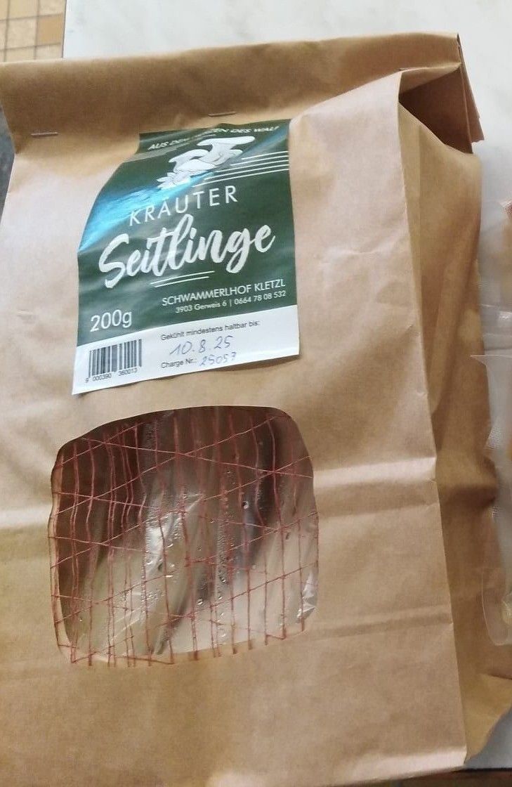 Ein brauner Papierbeutel mit einem Fenster, das Fische zeigt. Ein Etikett auf dem Beutel trägt 'Krauter Settlinge' und '200g'. Das Etikett enthält auch einen Barcode und zusätzlichen Text.