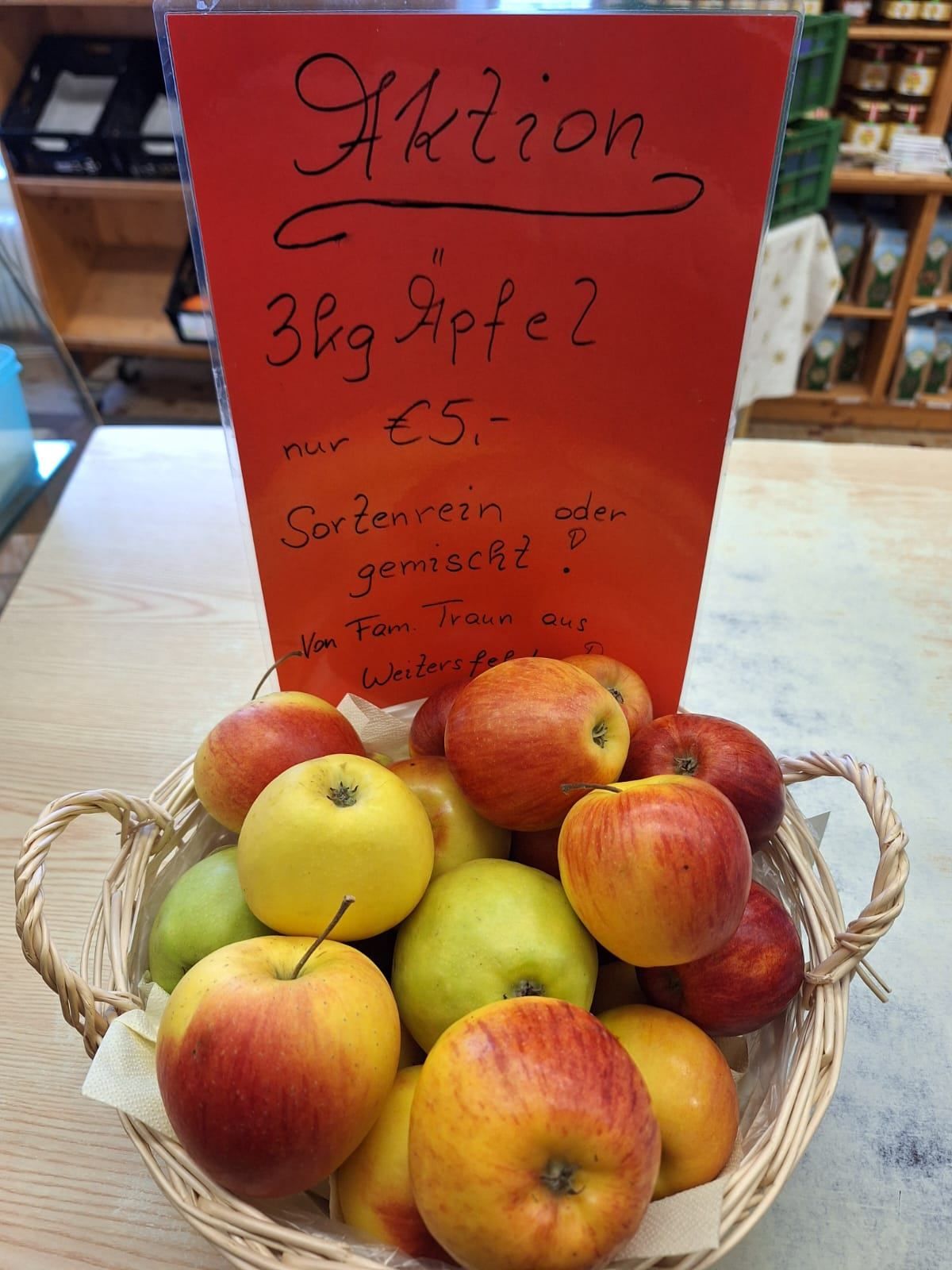 Ein Korb voller verschiedener Äpfel, darunter rot, gelb und grün, mit einem roten Schild darüber, das '3kg Äpfel für 5 Euro' steht. Die Äpfel liegen auf einem Holztisch.